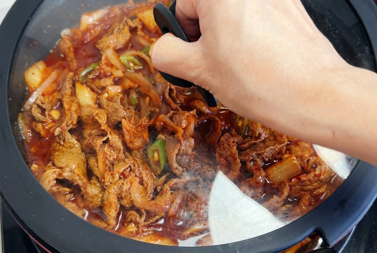 Une main posant un couvercle en verre sur une poêle où mijote le bœuf au kimchi dans une sauce rouge.