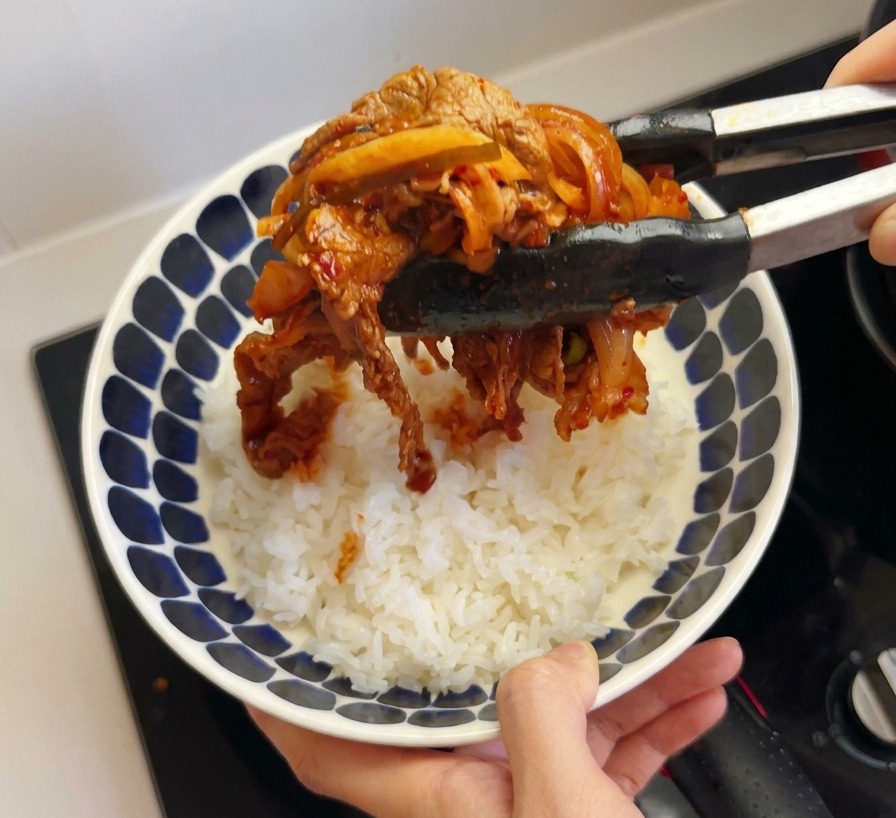 Des pinces de cuisine déposant le bœuf au kimchi sur un bol de riz blanc cuit.
