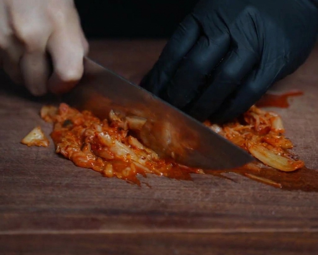 Gros plan dun chef hachant finement du kimchi rouge fermenté sur une planche en bois sombre avec un couteau bien aiguisé.