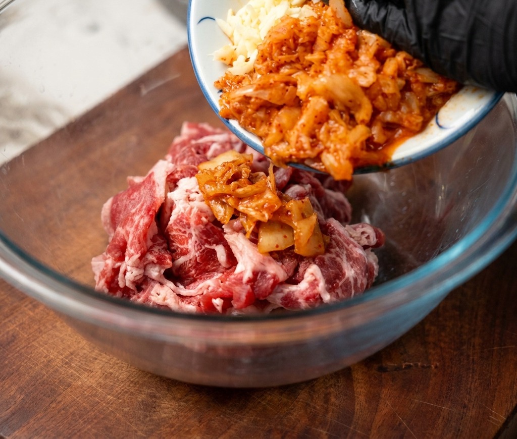 Du kimchi haché et de lail émincé versés depuis une petite assiette en céramique dans un bol contenant du bœuf cru finement tranché.