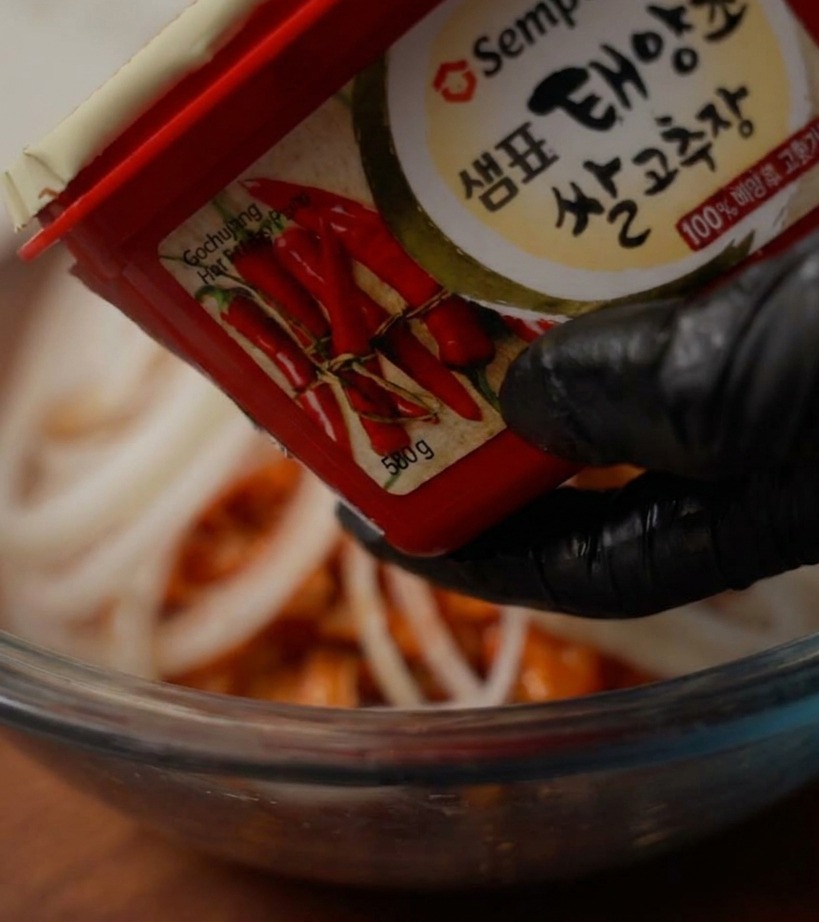 Une main gantée tenant un pot de pâte de piment rouge au-dessus dun bol pour y ajouter une cuillerée de pâte.