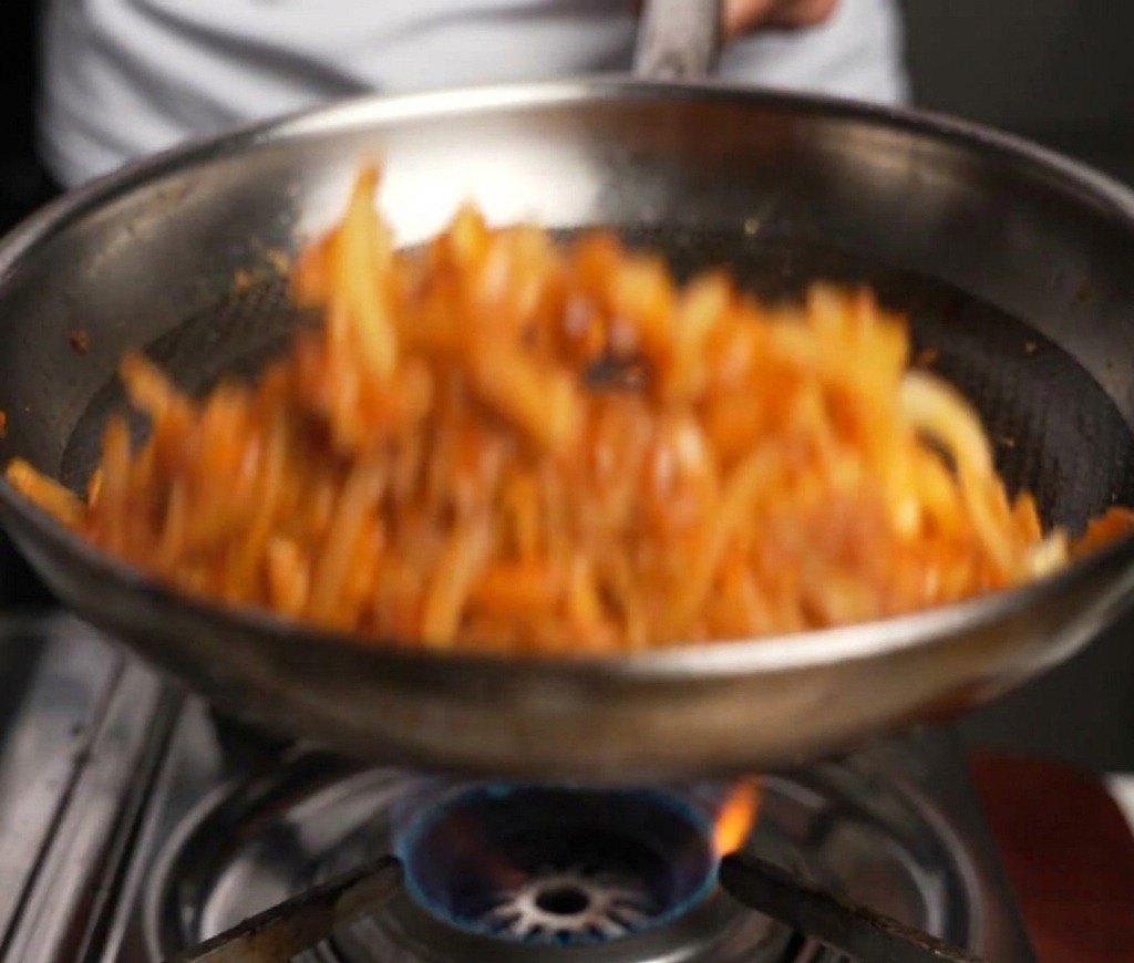 Bœuf et oignons remués dans une poêle métallique avec une spatule en bois sur une flamme de gaz.