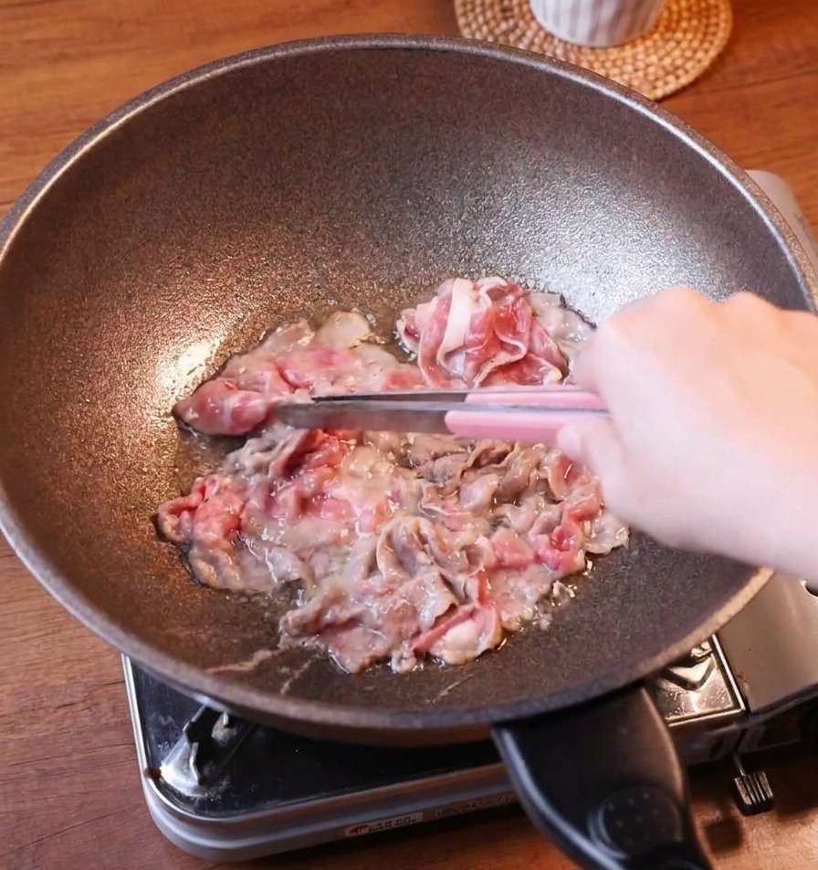 De fines tranches de bœuf doré retirées dune poêle en fonte à laide de pinces métalliques à manche rose.