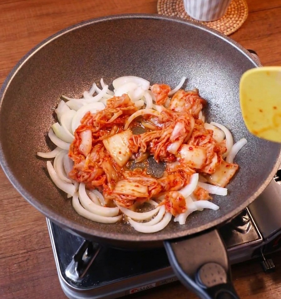 Des oignons émincés et du kimchi rouge vif sautés ensemble dans une poêle avec une spatule jaune.