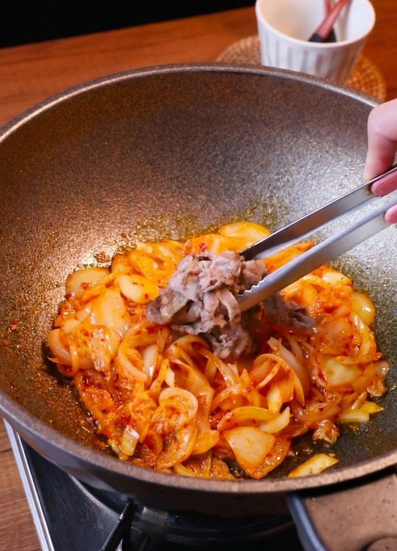 Une pince réintroduisant les tranches de bœuf doré dans la poêle remplie de kimchi et doignons fondus.
