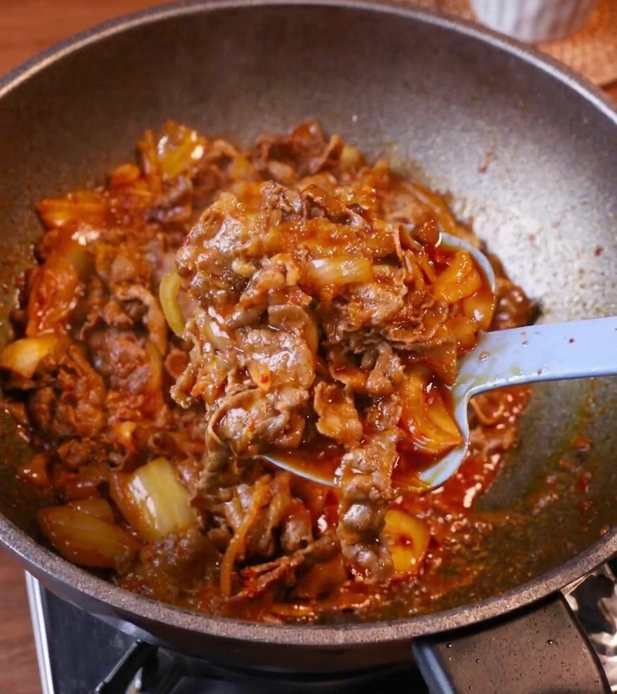 Une cuillère bleu clair déposant une portion généreuse de bœuf au kimchi glacé sur un bol de riz blanc fumant.