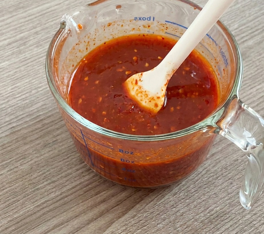Une spatule en silicone mélangeant une sauce rouge épaisse et brillante dans un verre doseur.