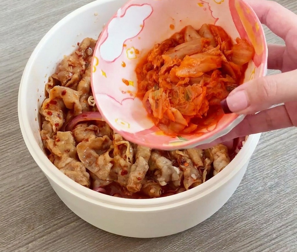 Une main versant un petit bol de kimchi rouge émincé dans une poêle contenant le bœuf et les oignons.