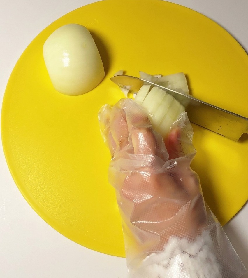 Une main gantée coupant un oignon blanc en petits dés avec un couteau en acier sur une planche à découper jaune.