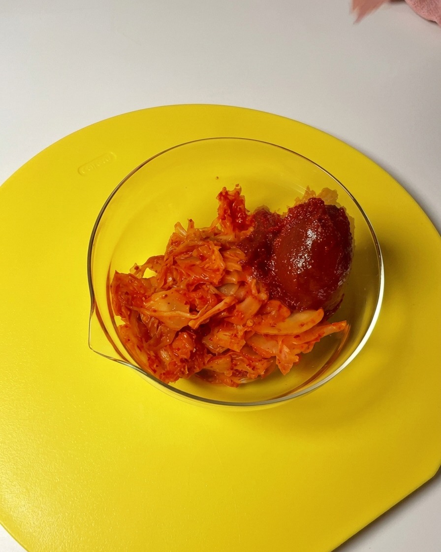 Une cuillerée de gochujang et de piment ajoutée à un bol en verre contenant du kimchi haché.