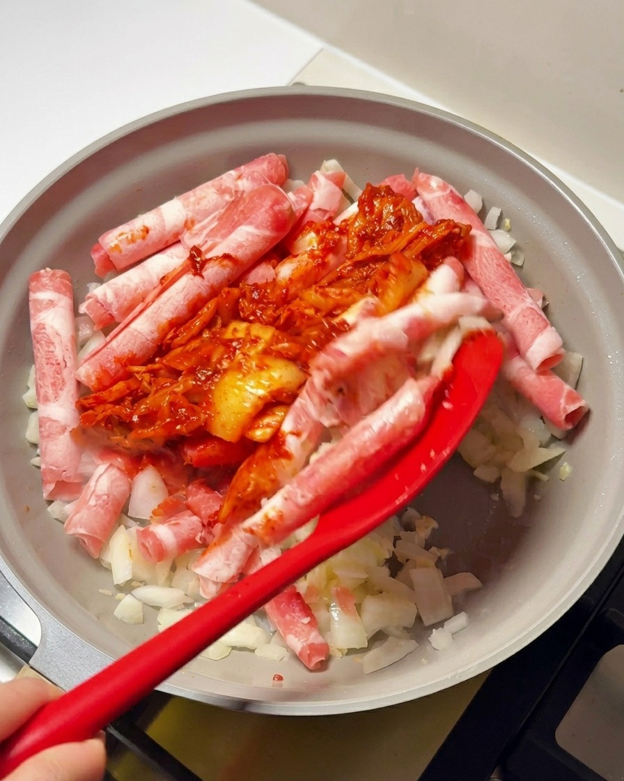 Spatule rouge ajoutant le mélange de kimchi épicé sur le bœuf dans la poêle.