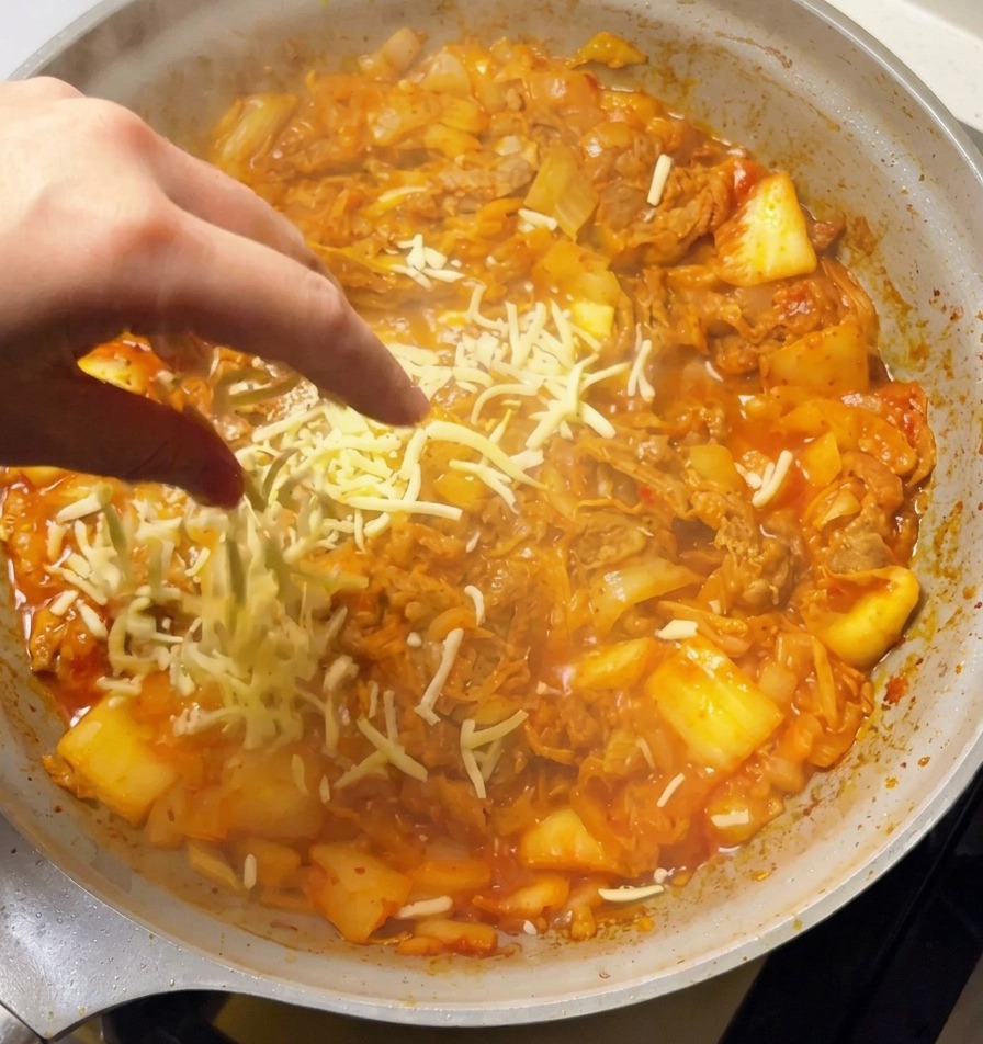 Main saupoudrant de la mozzarella râpée sur le mélange bouillonnant de bœuf et kimchi.