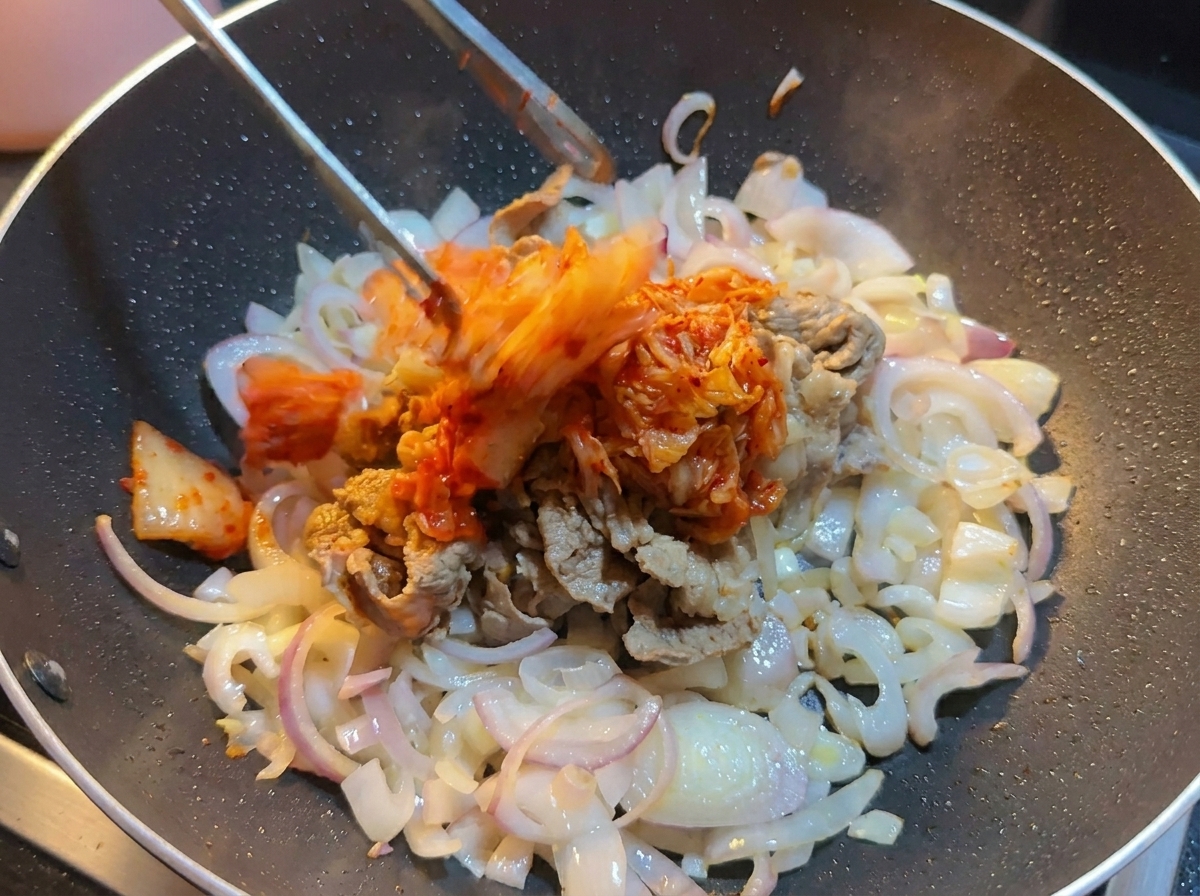 Kimchi de chou haché ajouté avec une pince dans un wok contenant le bœuf et les oignons.