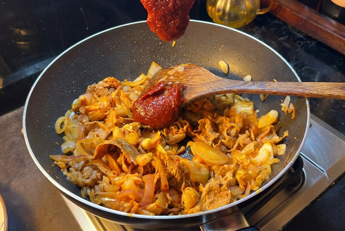 Une spatule en bois tenant une grosse dose de pâte de piment coréenne au-dessus de la poêle.