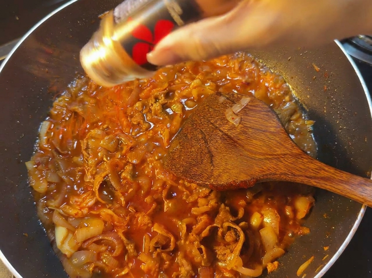 Poudre de piment rouge saupoudrée dans une poêle de sauce bouillonnante avec du bœuf et des oignons.