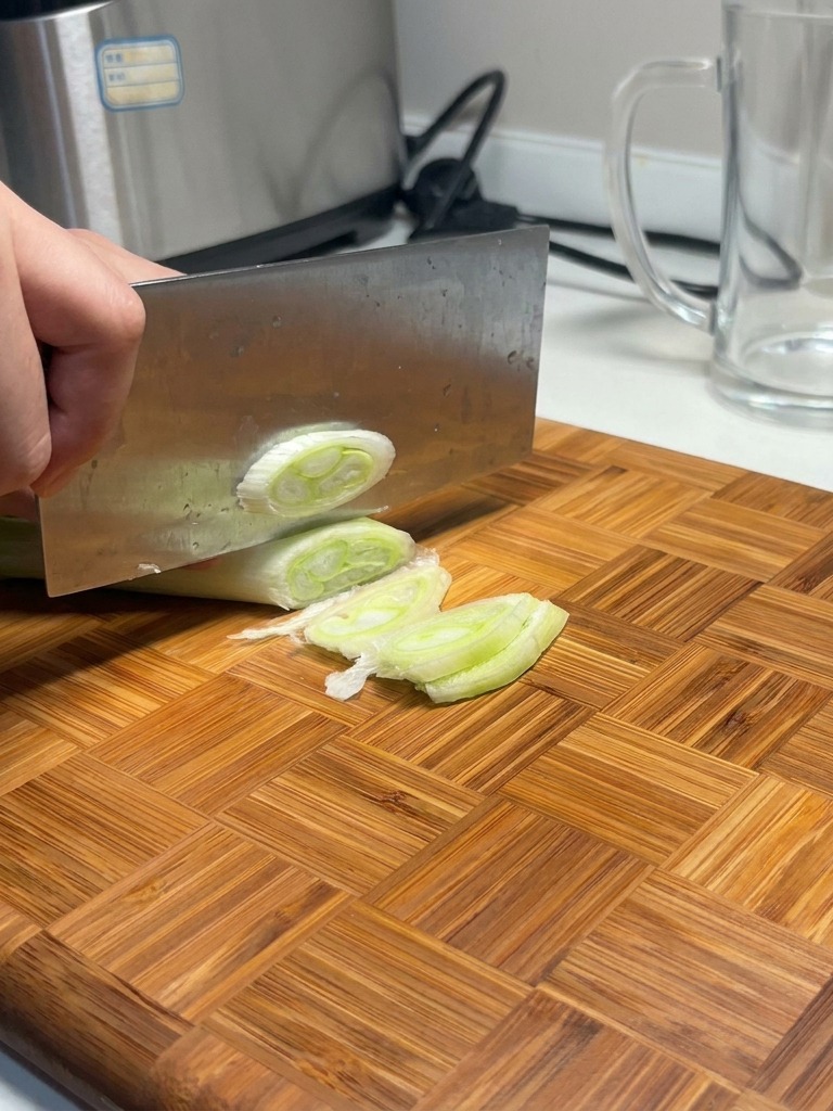 Un grand couperet tranchant un poireau épais en plusieurs rondelles blanches et vertes sur une planche en bois.