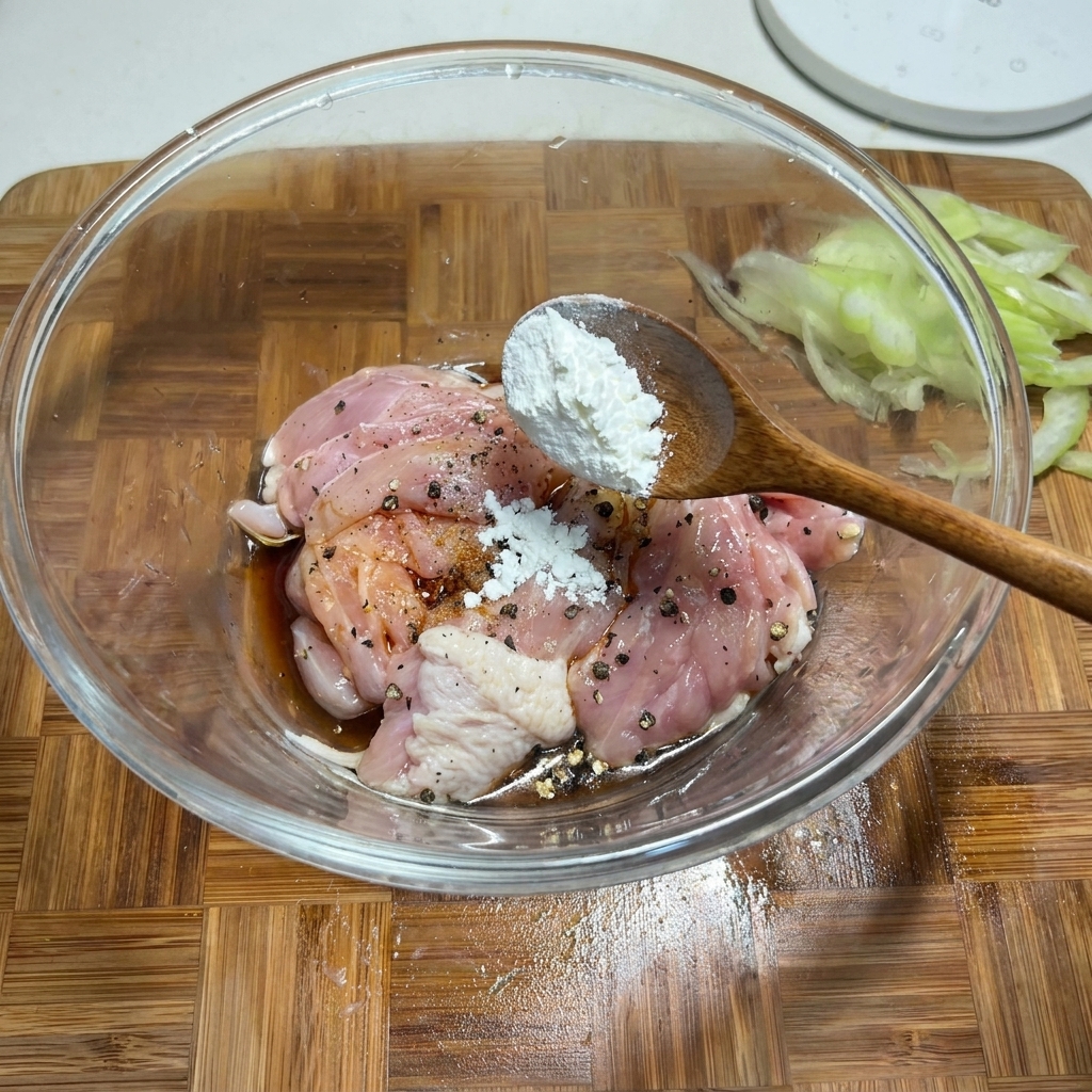 Une cuillère en bois ajoutant de la fécule de maïs blanche dans un bol en verre contenant des morceaux de poulet cru assaisonnés.