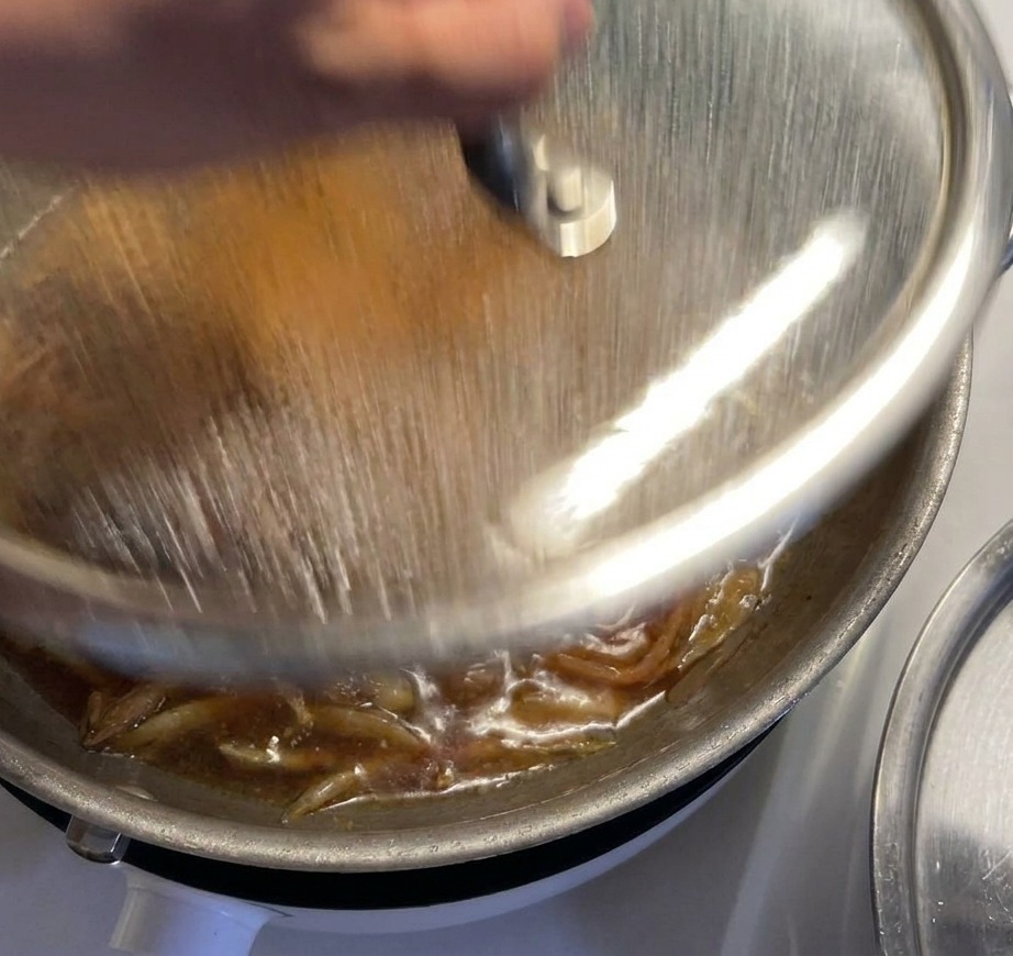 Une main posant un couvercle en verre sur une poêle où le poulet et les oignons mijotent dans une sauce bouillonnante.