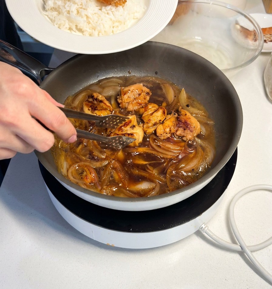 Des pinces métalliques déposant un morceau de poulet laqué sur un dôme de riz blanc dans un bol en céramique.