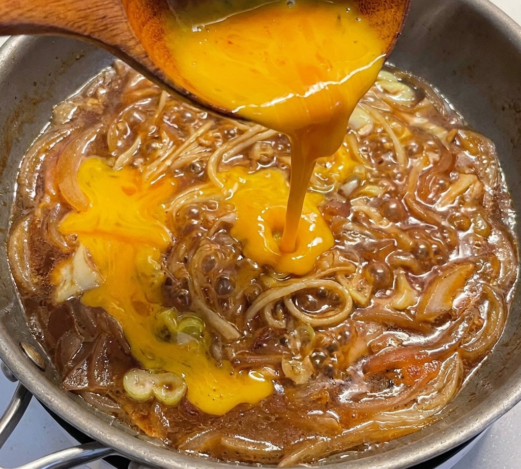 Œufs battus versés dune louche en bois dans une poêle remplie doignons et de poireaux mijotant dans la sauce.