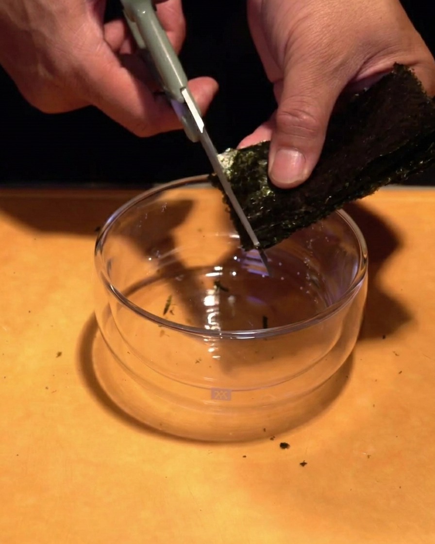Mains utilisant des ciseaux de cuisine pour couper une feuille dalgue vert foncé en fines lanières au-dessus dun bol en verre.