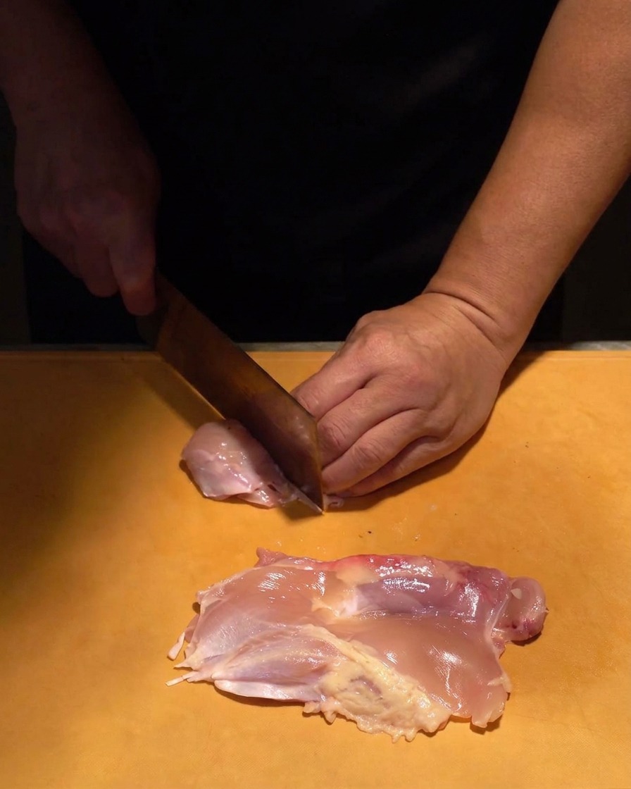 Viande de haut de cuisse de poulet crue et rosée coupée en morceaux uniformes avec un couteau bien aiguisé sur une planche orange.