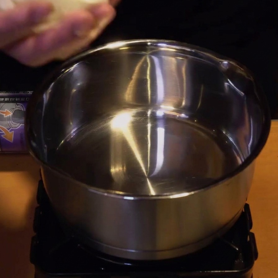 Une personne ajoutant des oignons émincés et des morceaux de poulet cru dans une petite casserole en inox sur un réchaud.