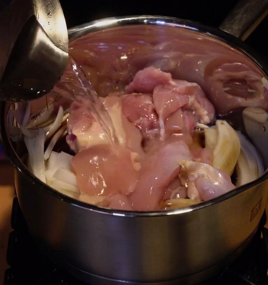 Eau versée dun pichet en métal dans une casserole remplie de hauts de cuisse de poulet et doignons émincés.