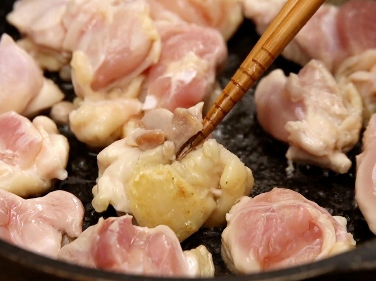 Dés de poulet dorant dans une poêle sombre, manipulés avec des baguettes.