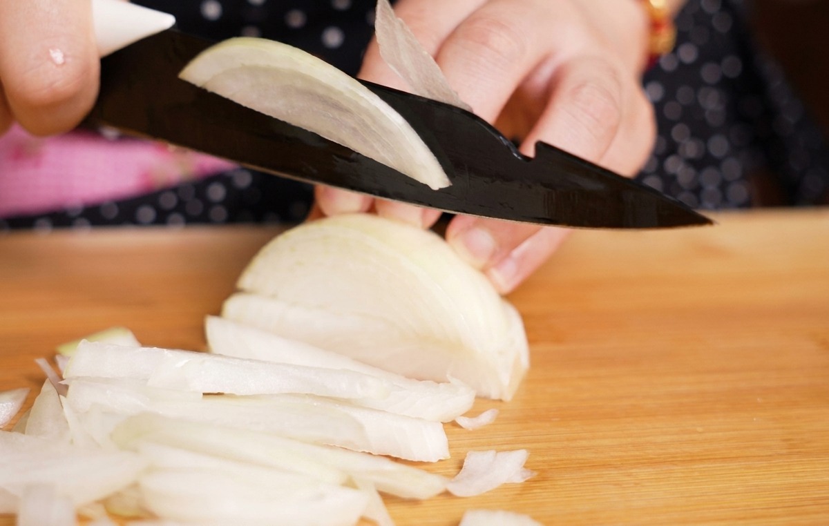 Une personne utilisant un couteau noir pour couper un oignon blanc en fines lanières sur une planche en bois clair.