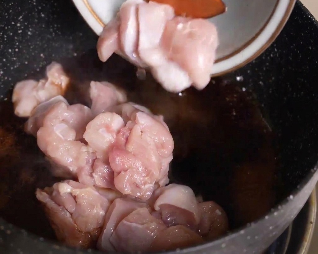 Morceaux de poulet cru ajoutés depuis un bol dans une poêle contenant une sauce sombre frémissante.