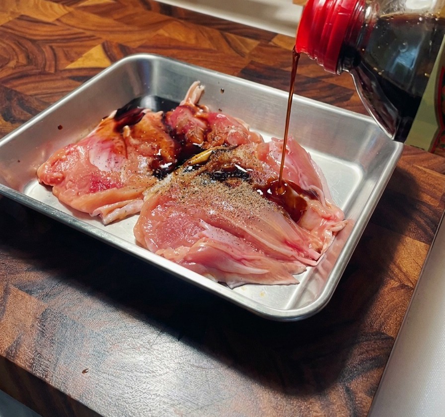 Sauce soja versée sur des hauts de cuisses de poulet crus et poivrés dans un plat en acier inoxydable.