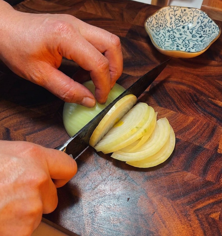 Mains dun chef utilisant un couteau pour couper un oignon jaune en fines lamelles sur une planche en bois sombre.