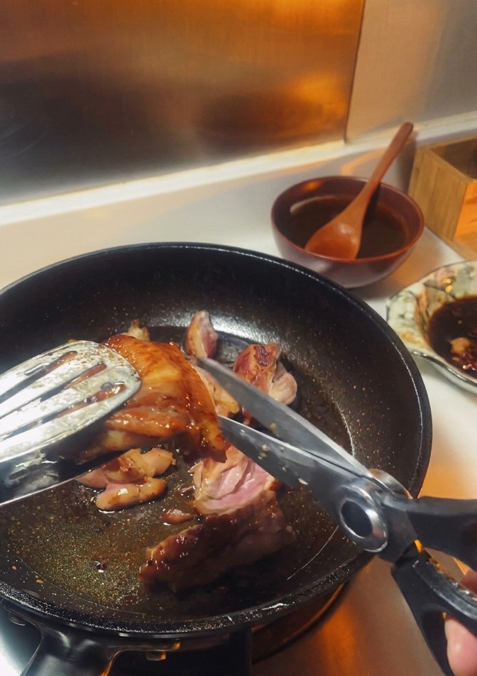 Ciseaux de cuisine découpant un haut de cuisse de poulet doré maintenu par des pinces directement dans la poêle.