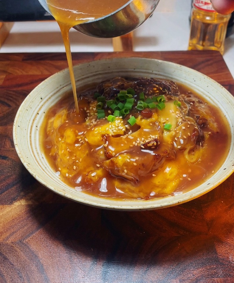 Gros plan dune main utilisant une louche en métal pour verser une sauce dorée sur un bol dOyakodon garni doignons verts.