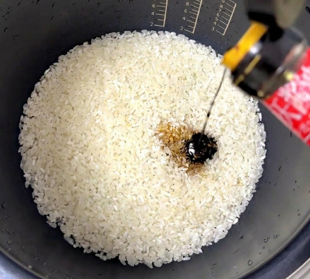Un filet de sauce dhuître épaisse et sombre versé dans un cuiseur à riz contenant du riz blanc lavé.