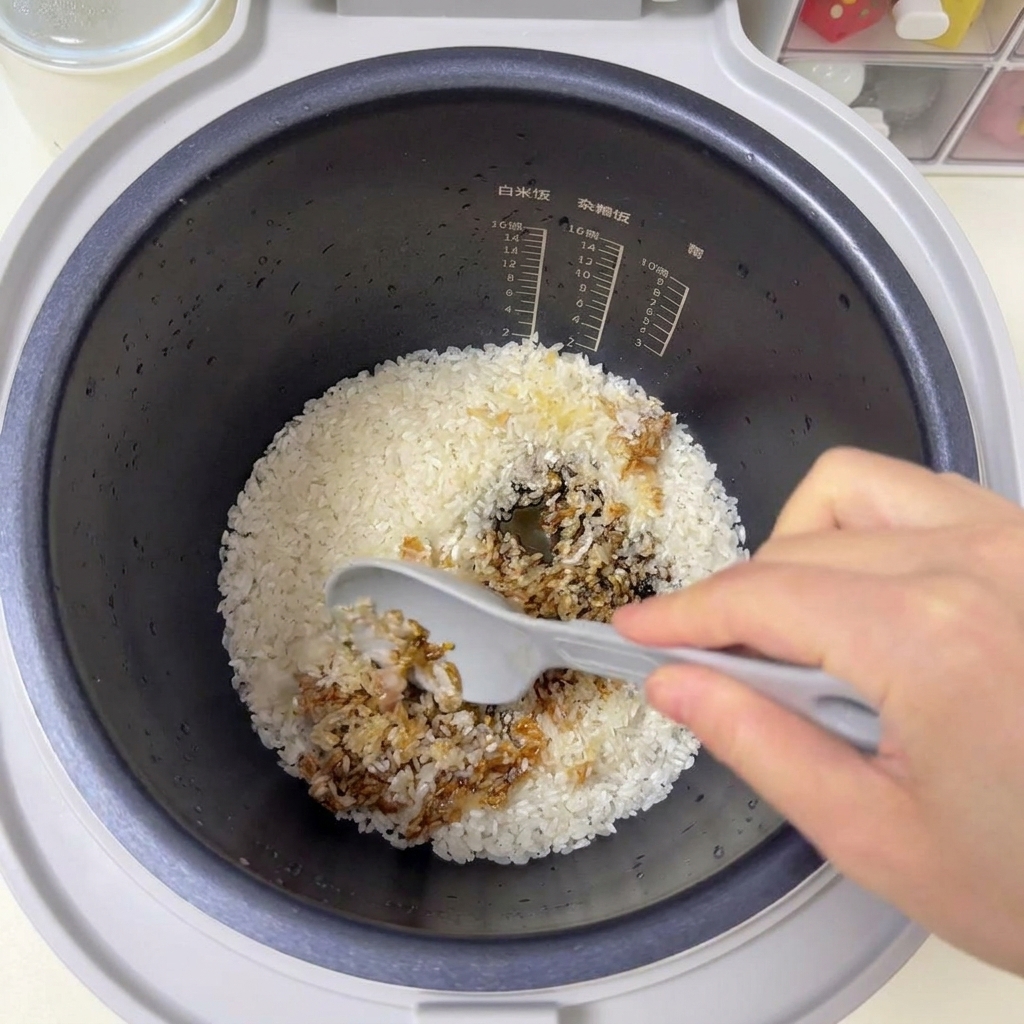 Une spatule grise mélangeant le riz blanc et les sauces brunes à lintérieur dun cuiseur à riz.