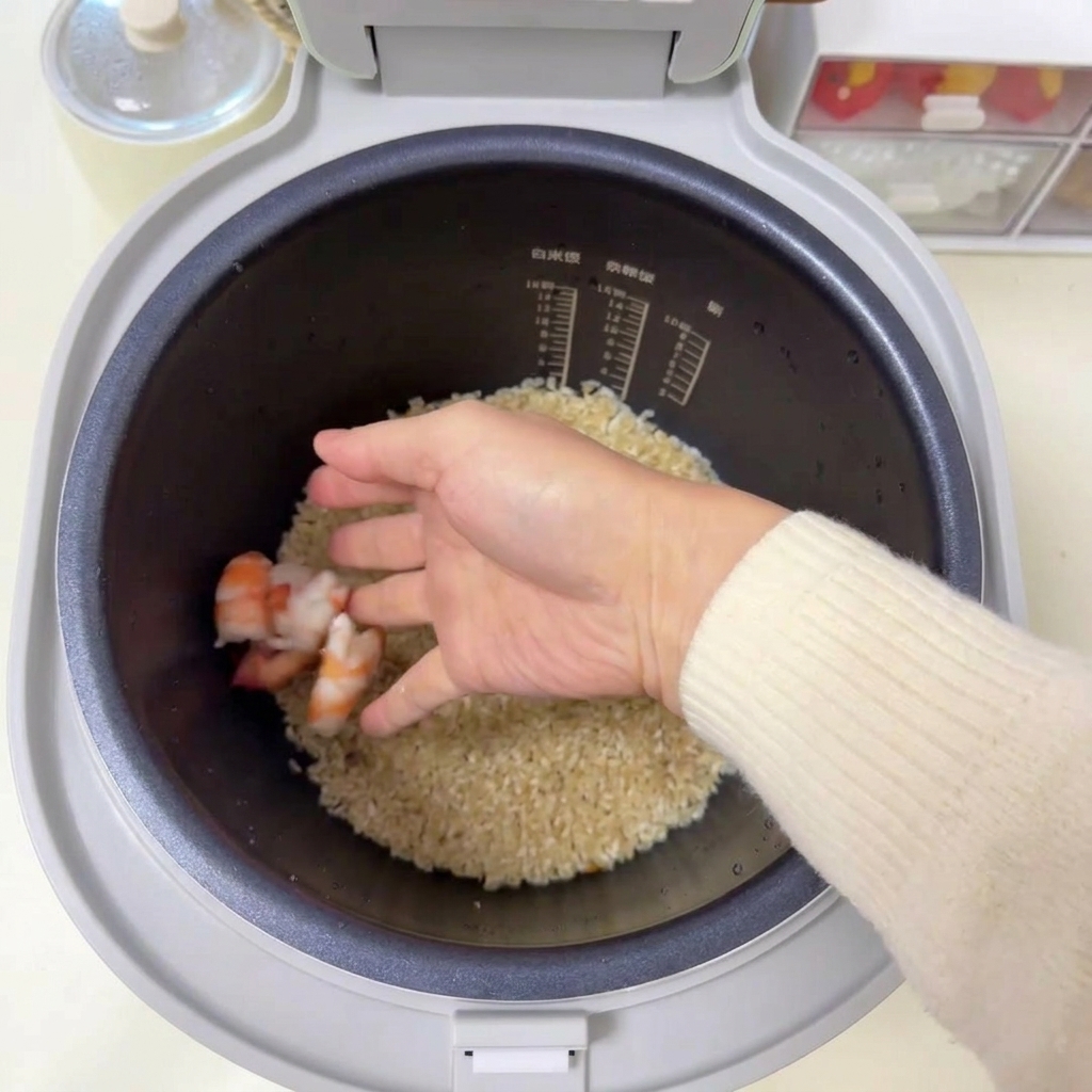 Une main plaçant plusieurs crevettes roses crues sur un lit de riz assaisonné dans un cuiseur à riz.