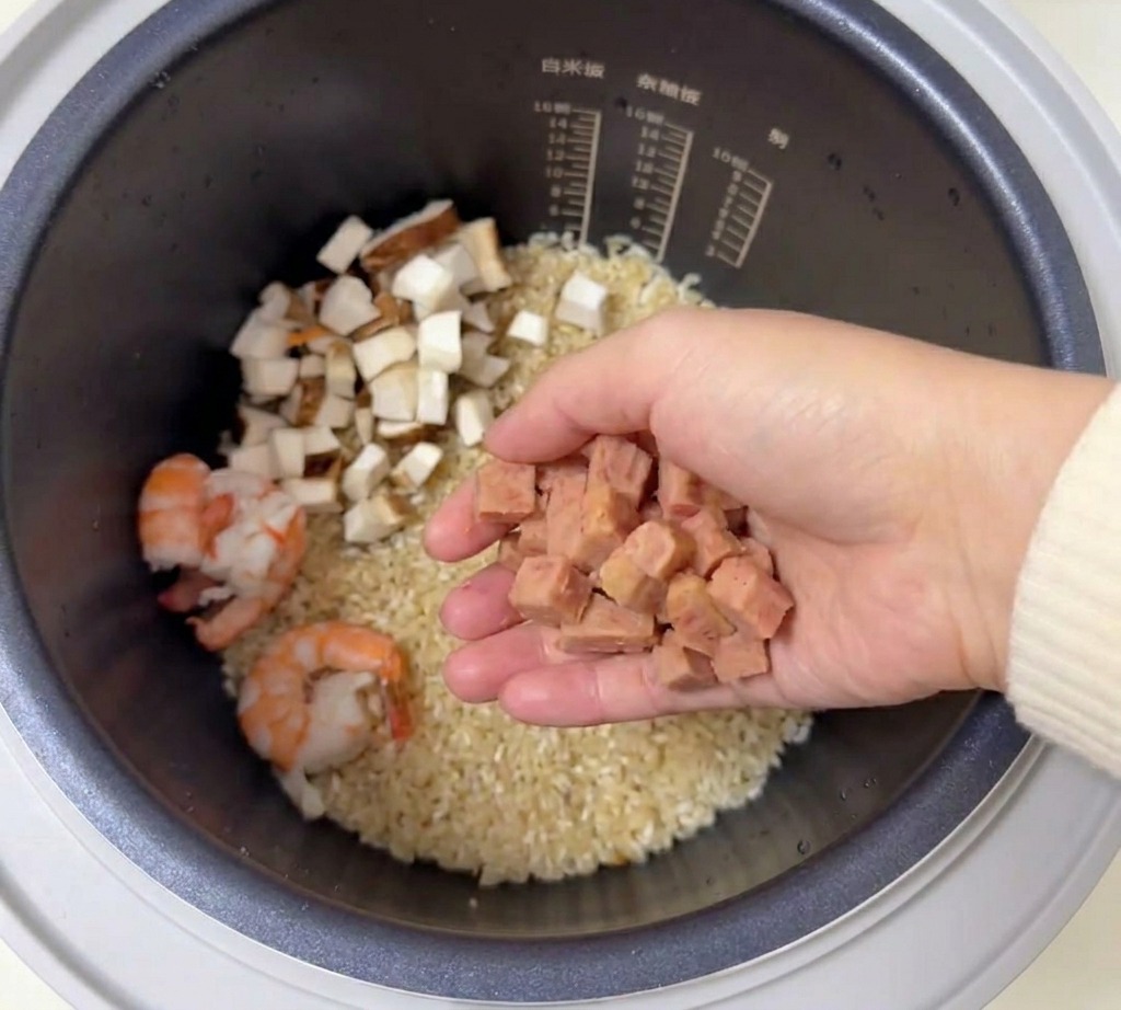 Une main tenant une poignée de dés de jambon rose au-dessus dun cuiseur à riz contenant du riz et des crevettes.