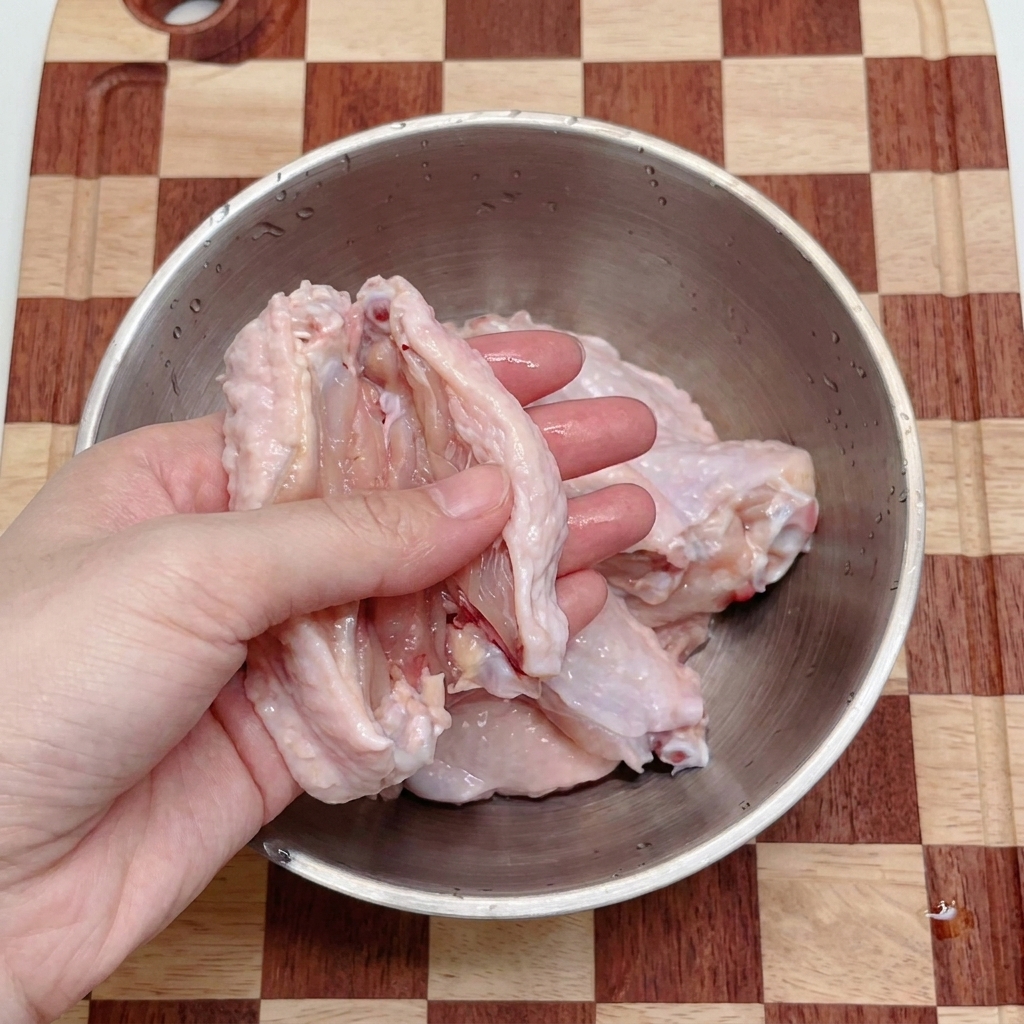 Une paire de mains utilisant des ciseaux de cuisine pour couper des ailes de poulet crues en deux dans un bol en inox.