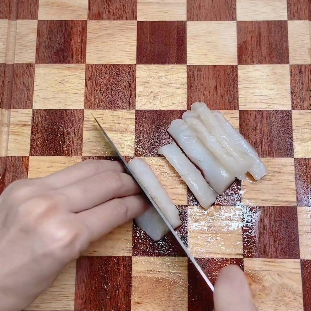 Un couteau de cuisine tranchant un filet de cabillaud blanc en lanières régulières sur une planche en bois.