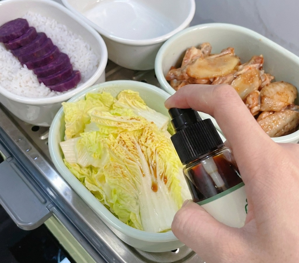 Une main utilisant un vaporisateur pour asperger de la sauce soja sur des feuilles de petit chou chinois dans un bol.