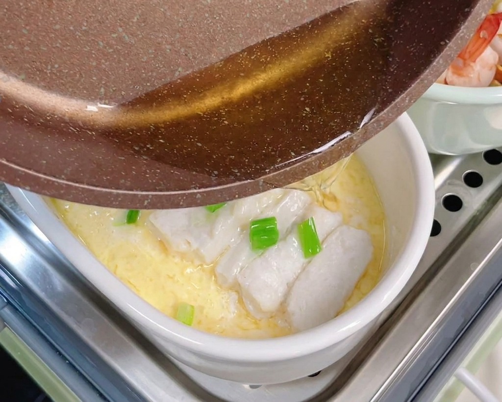 De lhuile chaude versée dune poêle sur des oignons verts hachés et du cabillaud vapeur reposant sur un flan dœufs.