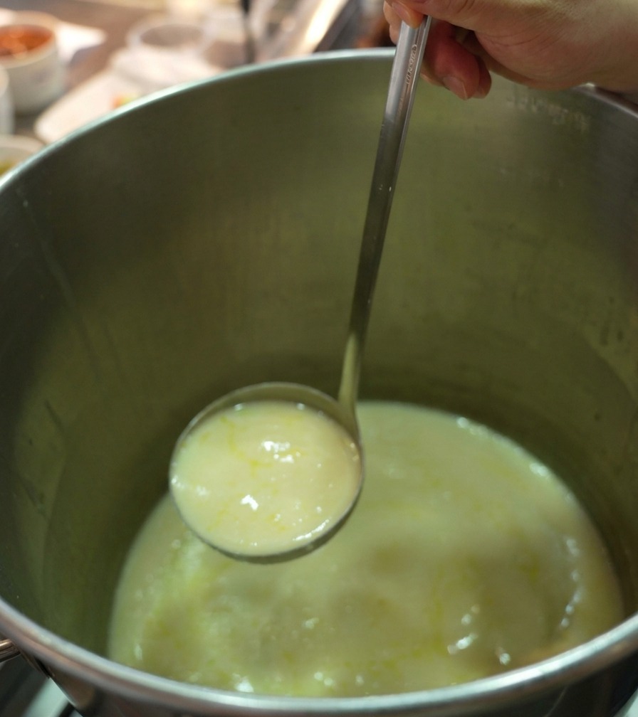 Une louche en acier inoxydable puisant un bouillon de poulet épais et crémeux dans une grande marmite professionnelle.