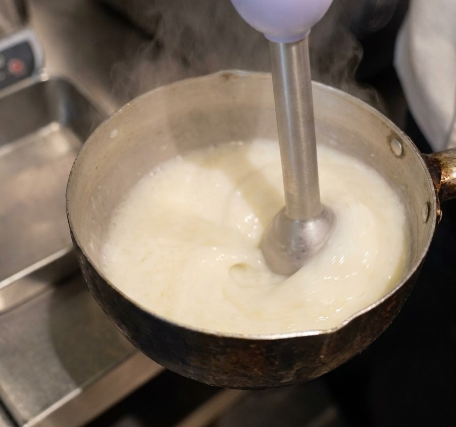 Un mixeur plongeant immergé dans un liquide jaune, créant une mousse épaisse et blanche à la surface.