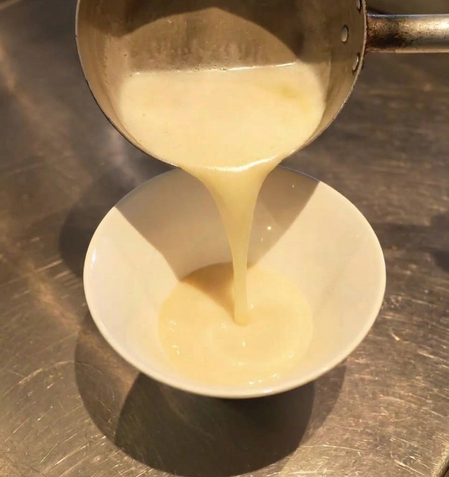 Un bouillon blanc laiteux versé dune casserole dans un bol blanc, se mélangeant à lassaisonnement sombre au fond.