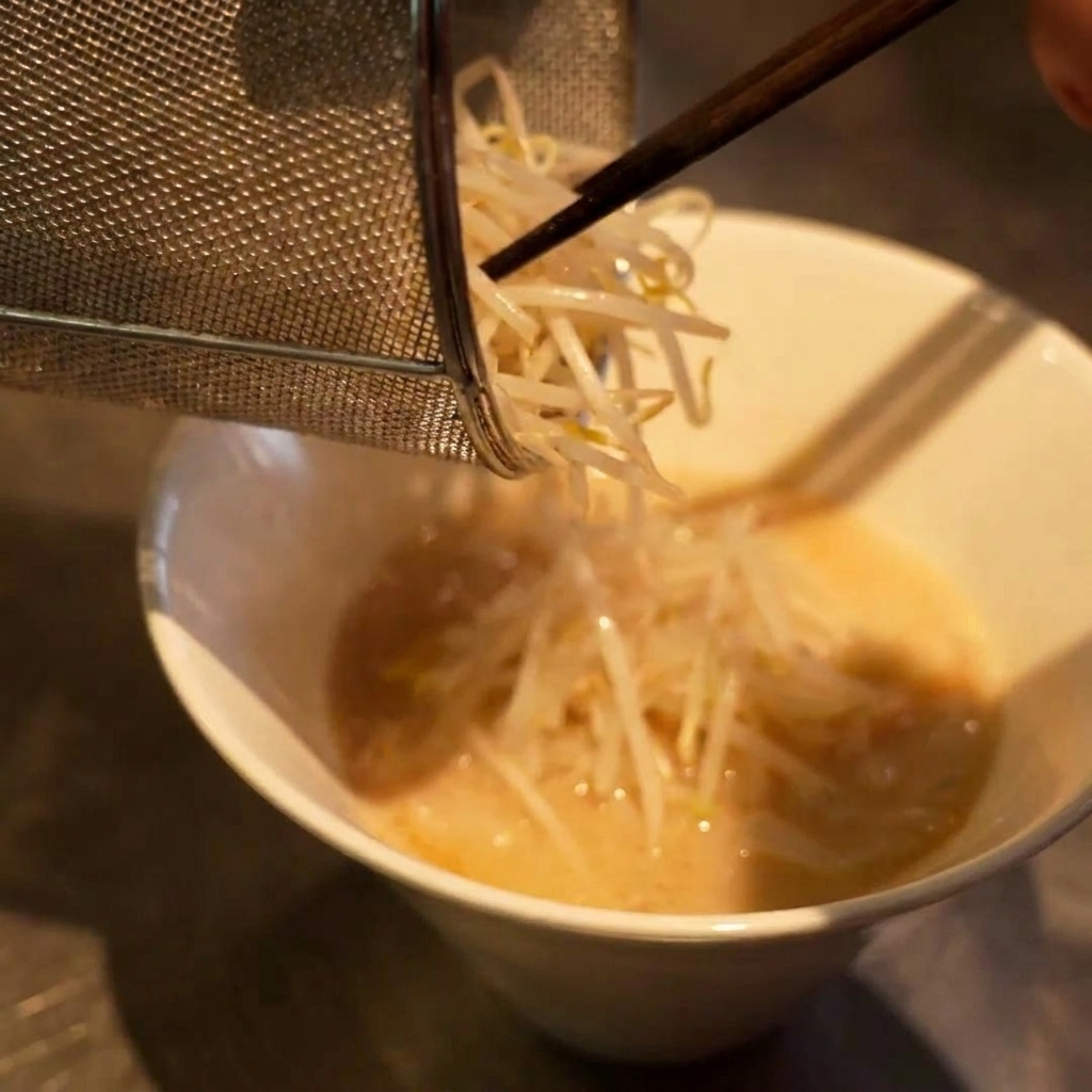 Une petite passoire déposant un tas de pousses de soja blanches sur un bol de ramen.