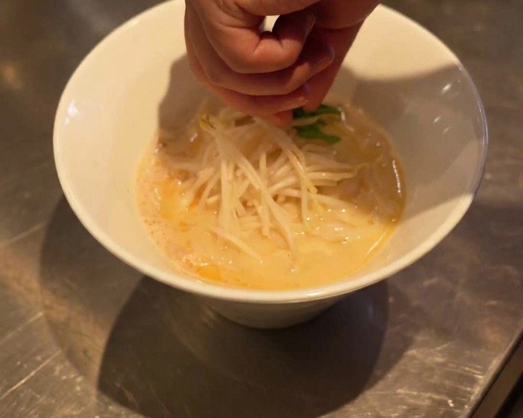 Une main plaçant avec précaution des feuilles dépinards frais sur des pousses de soja dans un bol de ramen.