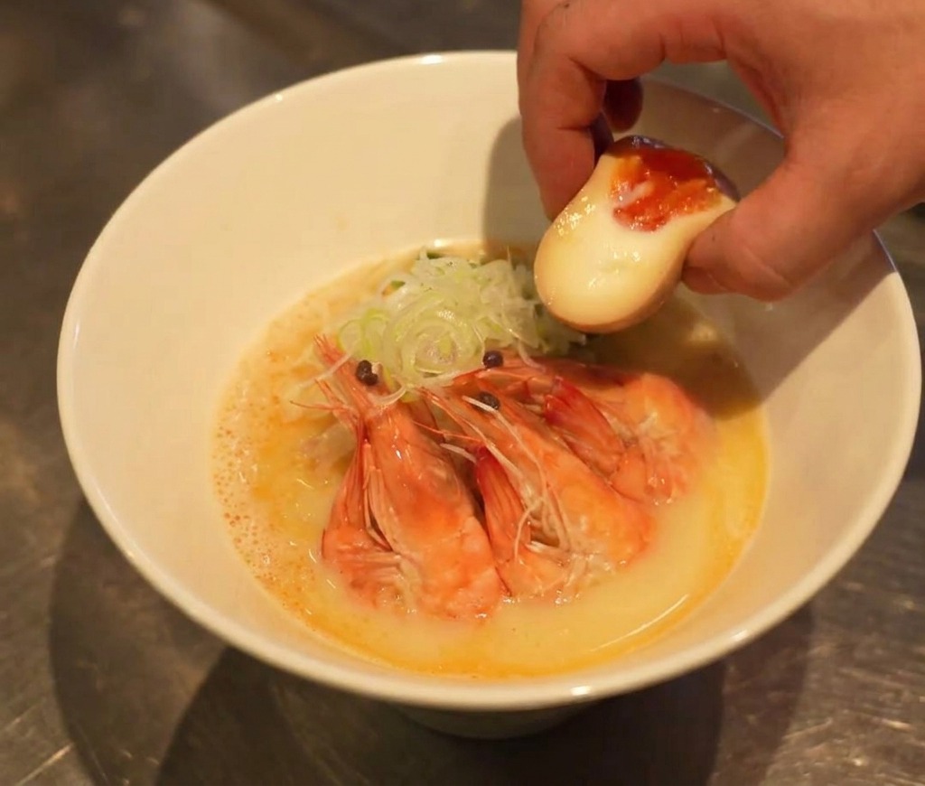 Une main plaçant une moitié dœuf mollet au jaune coulant dans un bol de ramen presque terminé.