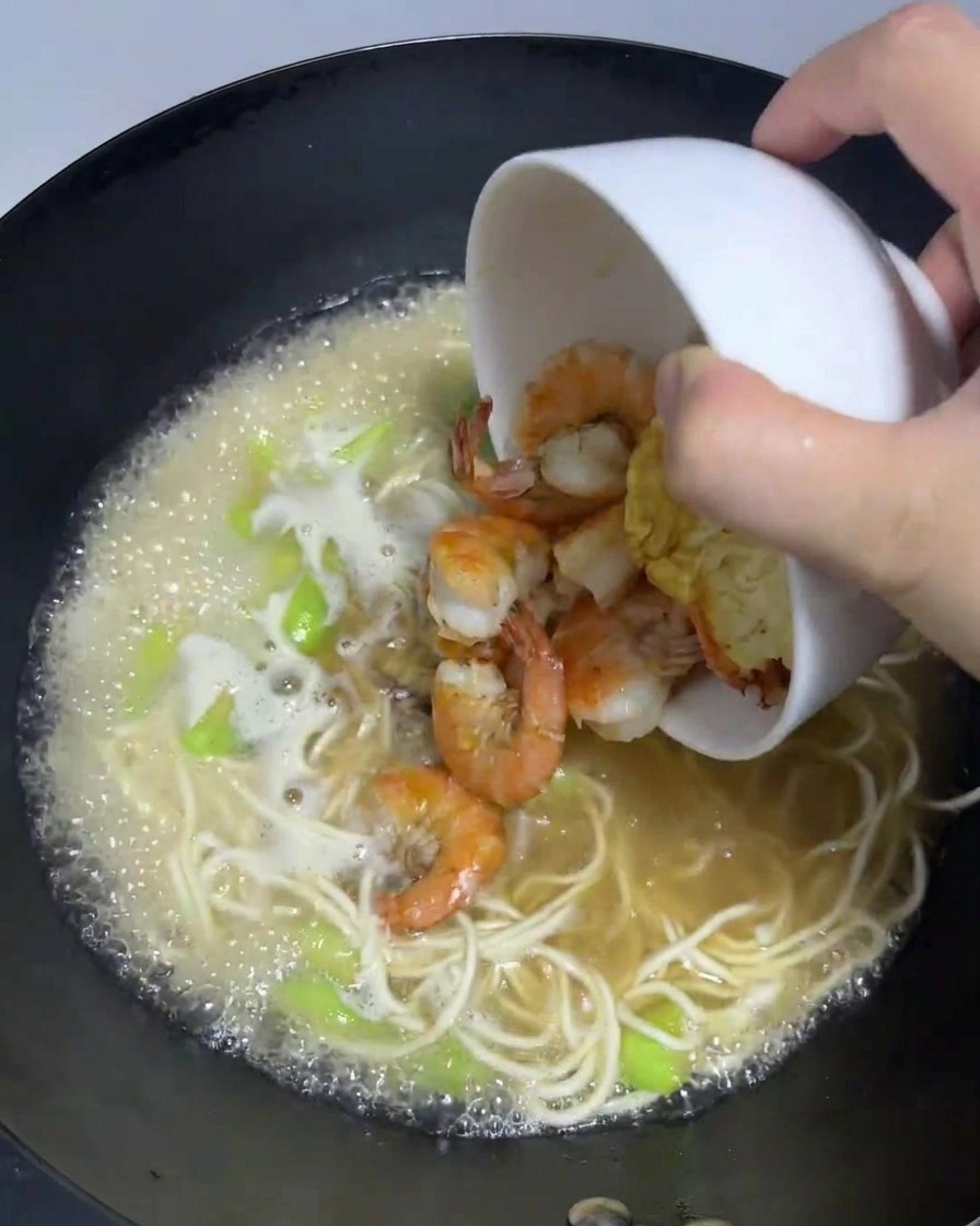 Crevettes cuites et œuf frit versés dans le wok de ramen frémissant.